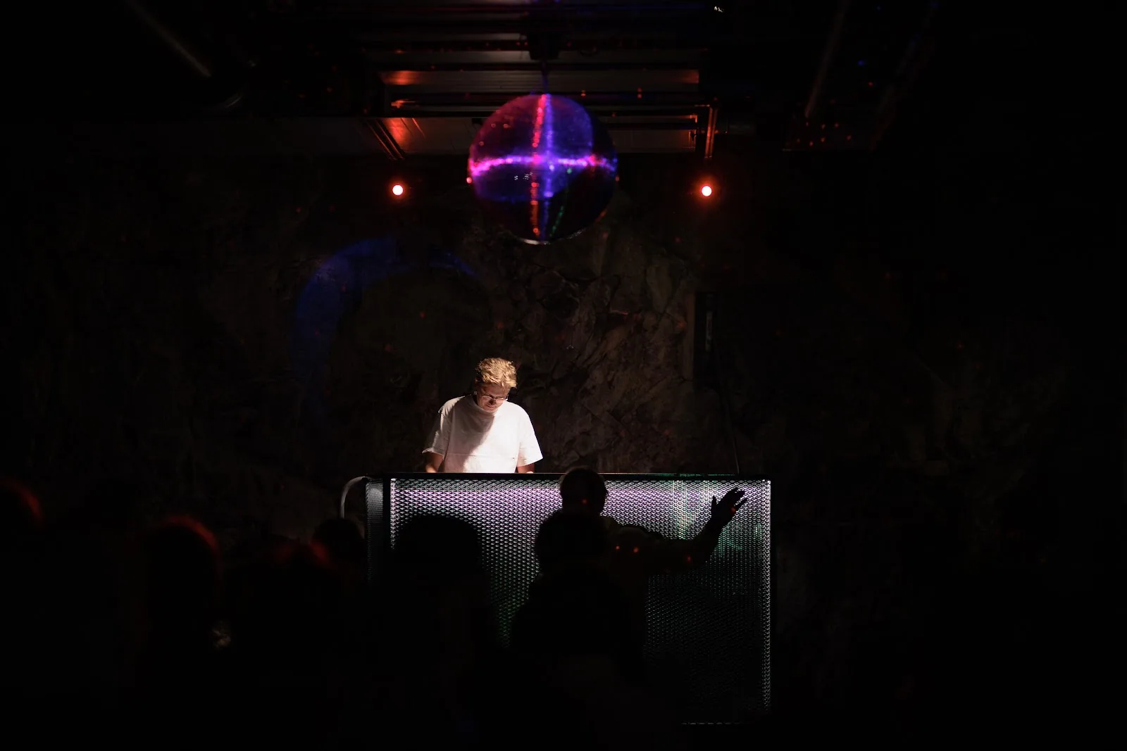 A DJ beneath a purple disco ball, with a raised hand silhouetted in front of the booth. Stockholm.