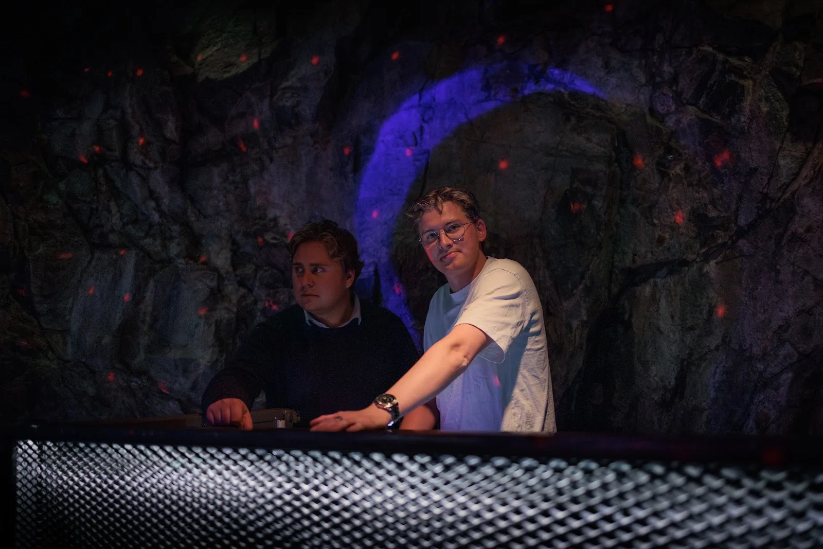 Two DJs behind the mesh booth against a rough rock wall and purple light. Stockholm.